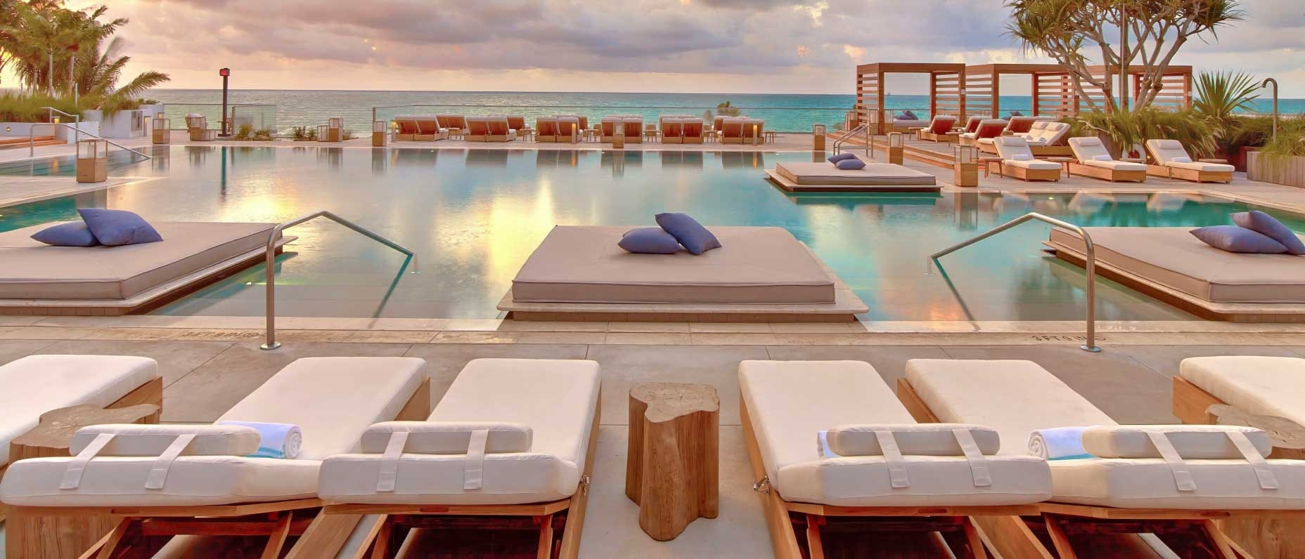 Vue d’une piscine entourée de lits de repos et de chaises longues, avec des palmiers et l’océan en arrière-plan.