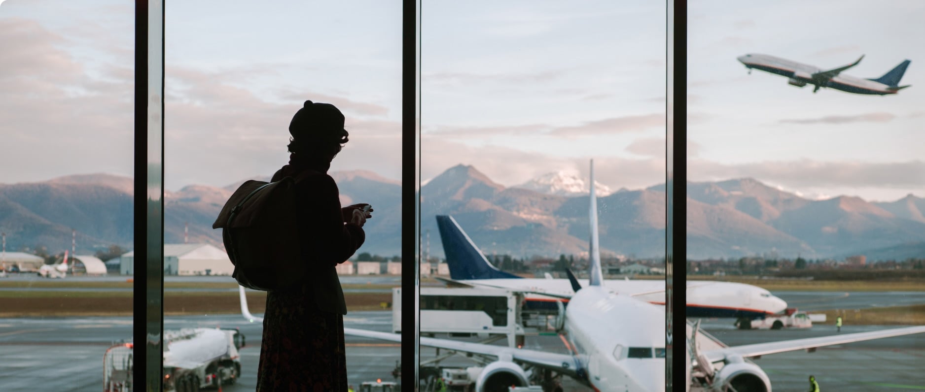 A traveler stands in an airport terminal looking out at the planes on the tarmac with mountains in the distance
