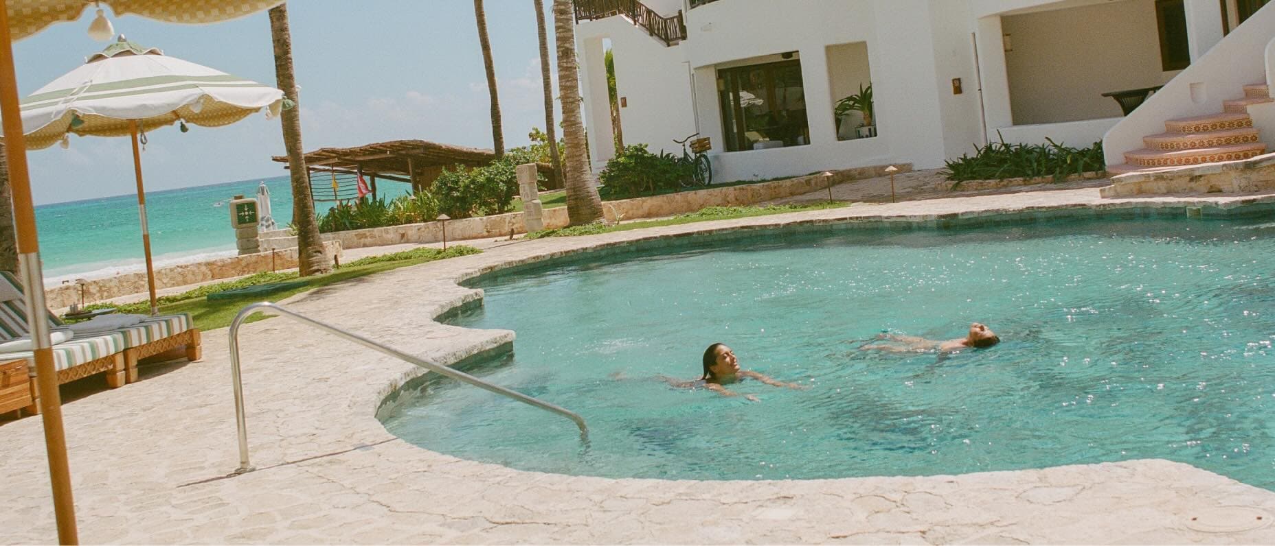 Two people swim in a hotel pool with aqua-blue ocean waters visible in the background