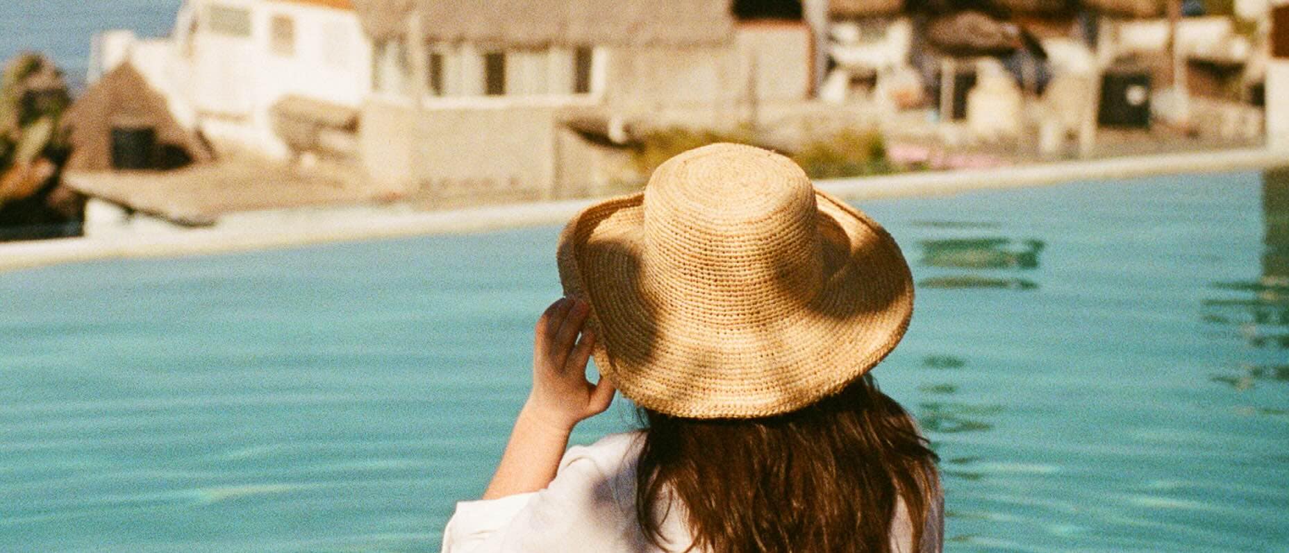 A woman with long brown hair and a straw hat dips her feet in an infinity pool as she looks out at the sea