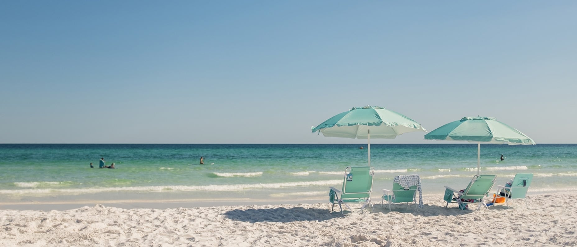 Turquoise beach umbrellas and lounge chairs on white sand, calm teal ocean with swimmers in the distance.