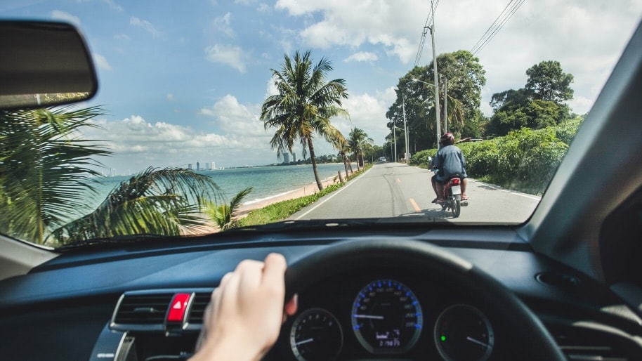 Driving along a coastal road, palm trees sway on the left with the blue ocean beyond. A person on a motorbike is ahead under the sunny sky.