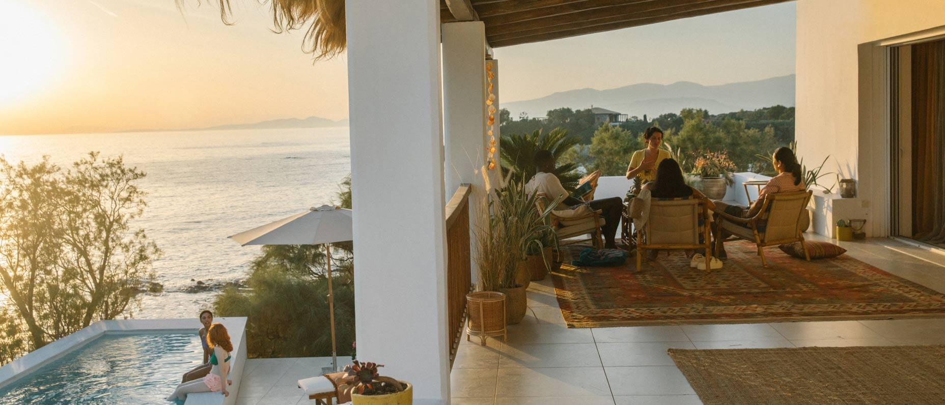 Ein Terrassenpool mit Sonnenliegen und -schirmen sowie Blick auf grüne Bäume und das Meer. Die Szenerie wirkt ruhig und einladend.