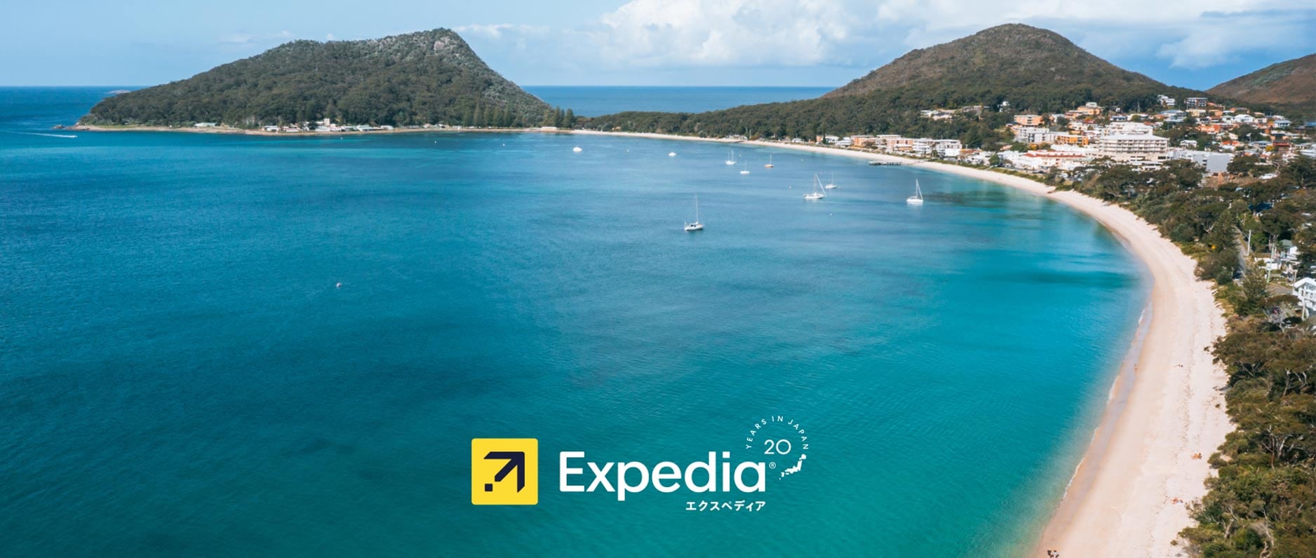 Aerial view of a curved white-sand beach and turquoise bay flanked by two tree-covered hills, with sailboats on the water and a coastal town visible on the right, overlaid with the Expedia logo and a '20 Years in Japan' anniversary badge.