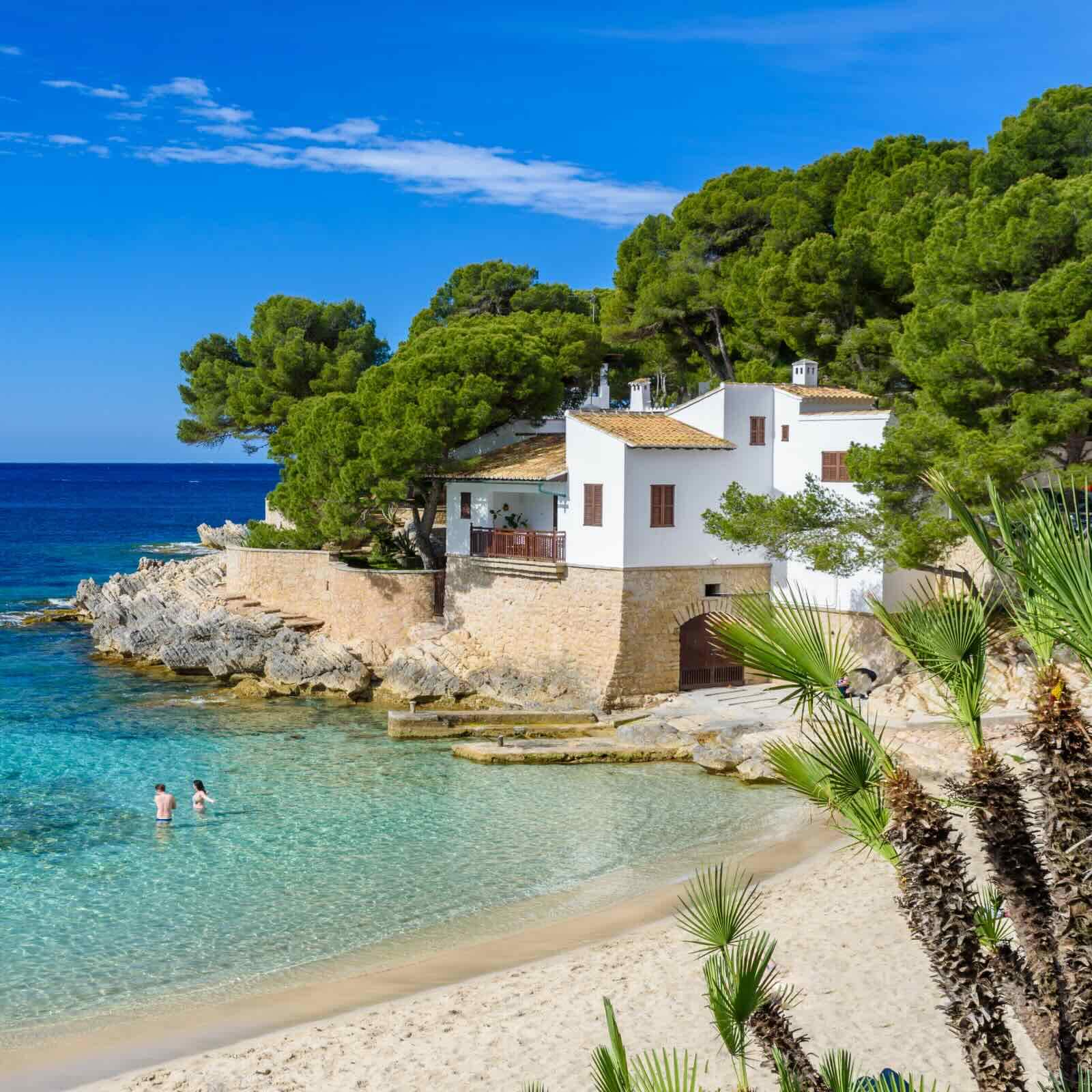 A coastal cove featuring a clear, turquoise bay and a white sandy beach. A white traditional house with wooden shutters is set above the rocky shoreline, with dense green trees in the background. Two people are wading in the shallow sea under a bright blue sky with light clouds. 