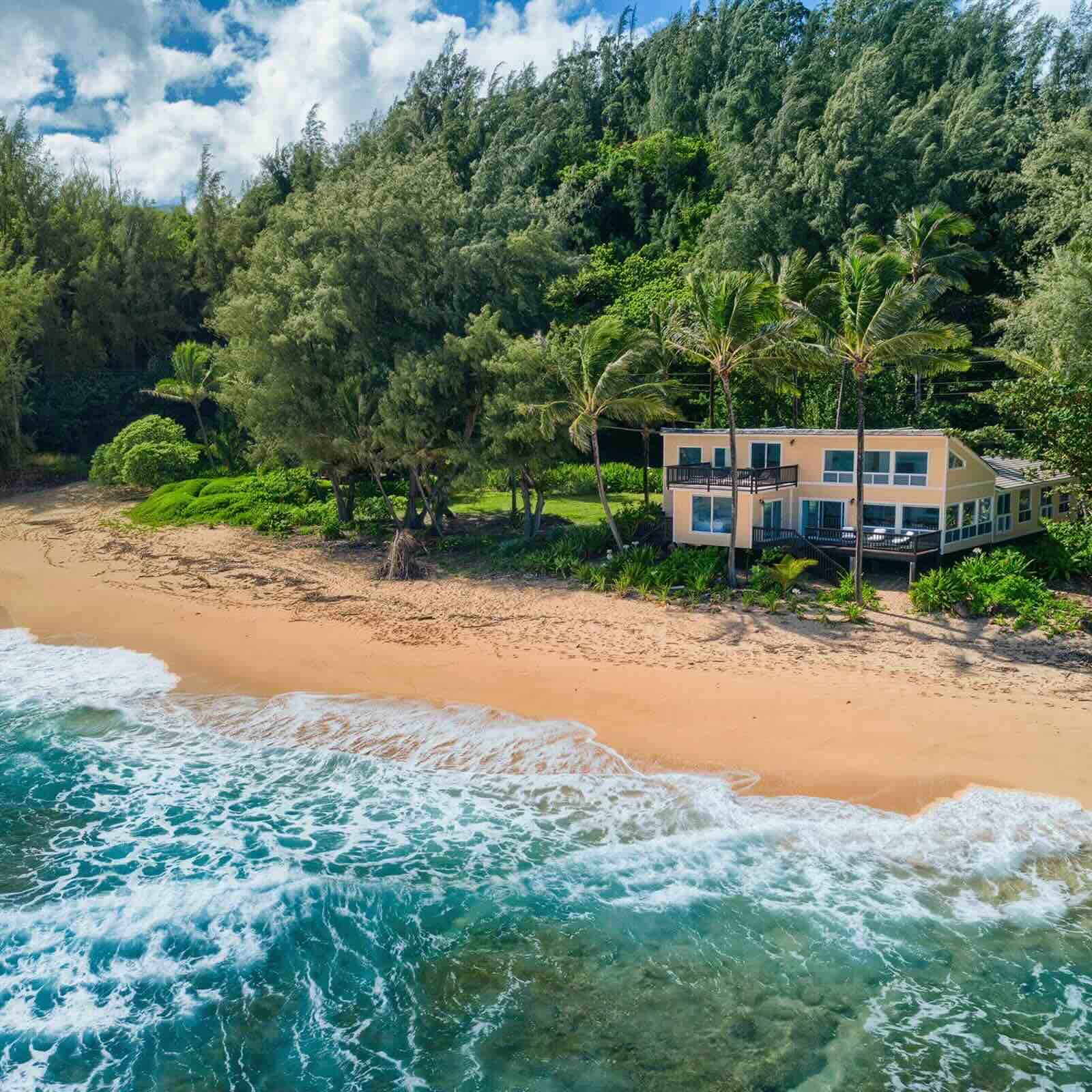 An aerial view of a two-story, yellow beachfront house on a secluded beach in Kauai, Hawaii. A dense green forest rises steeply behind the home, while palm trees stand in front. Turquoise ocean waves with white foam crash onto the golden sand beach in the foreground under a partly cloudy sky. 