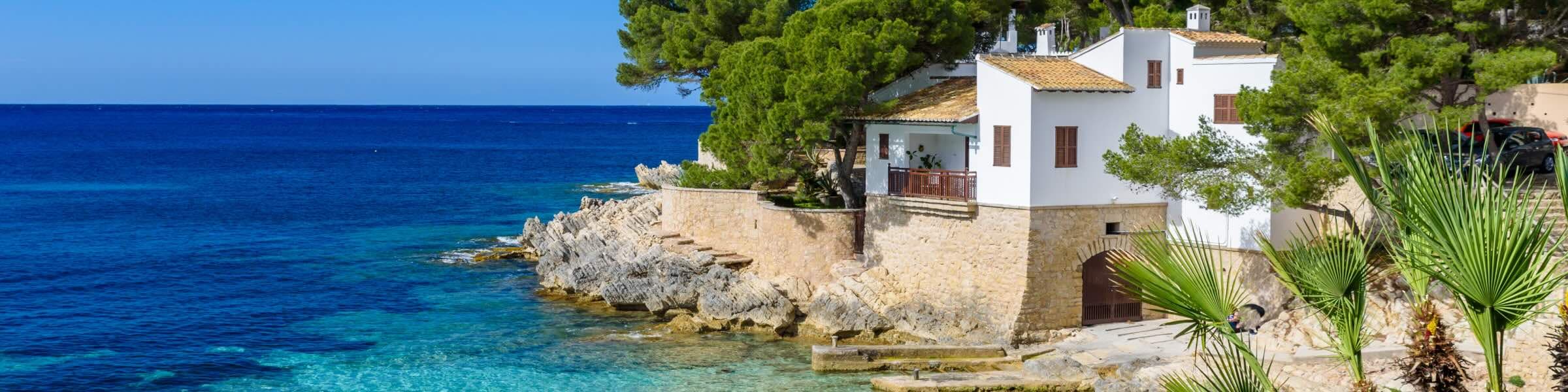 Une crique abrite une baie aux eaux turquoise cristallines et une plage de sable blanc. Une maison traditionnelle blanche aux volets en bois surplombe le rivage, avec une végétation dense à l’arrière-plan. Deux personnes se baignent dans la mer peu profonde sous un ciel d’un bleu éclatant parsemé de quelques nuages. 