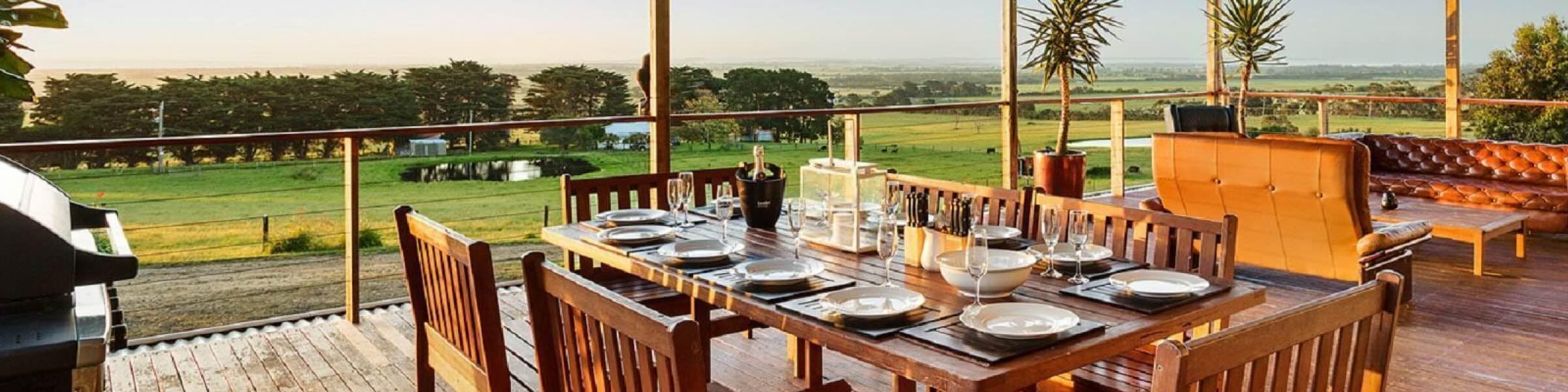 A spacious, covered wooden deck overlooks tranquil green fields and lakes at sunset. A large wooden dining table in the foreground is set for an evening meal, with a barbecue behind. In the background, a cosy outdoor lounge area features a two-seater couch, armchair and Chesterfield sofa. 