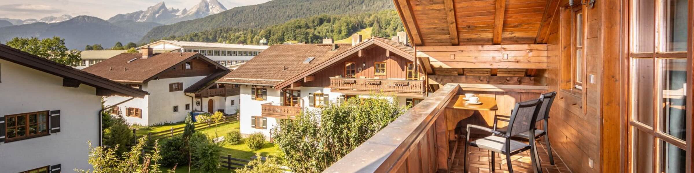 Ein sonniger Holzbalkon eines rustikalen Chalets blickt auf ein Tal mit traditionellen Häusern, dunklen Dächern und grünen Gärten. Im Hintergrund erheben sich Alpengipfel hinter dicht bewaldeten Hügeln. Der Balkon hat Terrakottafliesen, drei schwarze Netzstühle und einen kleinen Tisch mit zwei Teetassen und Untertassen. 
