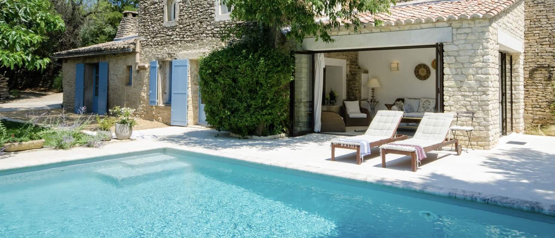 Vue panoramique d’une villa en pierre en Provence, par une journée claire et ensoleillée. Une piscine d’un bleu éclatant scintille au premier plan, tandis que deux chaises longues blanches sont disposées sur une terrasse en pierre claire au bord de l’eau. Un coin salon doté de baies vitrées s’ouvre sur la façade de la maison, laissant entrevoir des sièges confortables à l’intérieur.