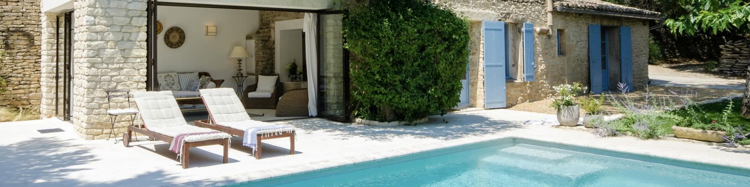 Vue panoramique d’une villa en pierre en Provence, par une journée claire et ensoleillée. Une piscine d’un bleu éclatant scintille au premier plan, tandis que deux chaises longues blanches sont disposées sur une terrasse en pierre claire au bord de l’eau. Un coin salon doté de baies vitrées s’ouvre sur la façade de la maison, laissant entrevoir des sièges confortables à l’intérieur.