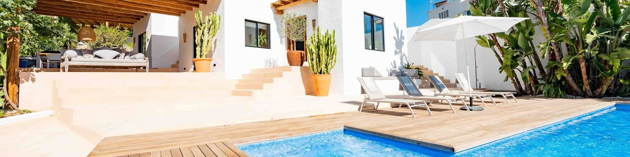 Under the clear blue Ibizan skies, a bright white villa is positioned towards the back of the view. To the left of the villa, a rustic wooden pergola covers an outdoor lounge area, surrounded by pots of large cacti. In the foreground, wooden decking surrounds a turquoise pool, with four sun loungers and a white umbrella positioned on the decking. 