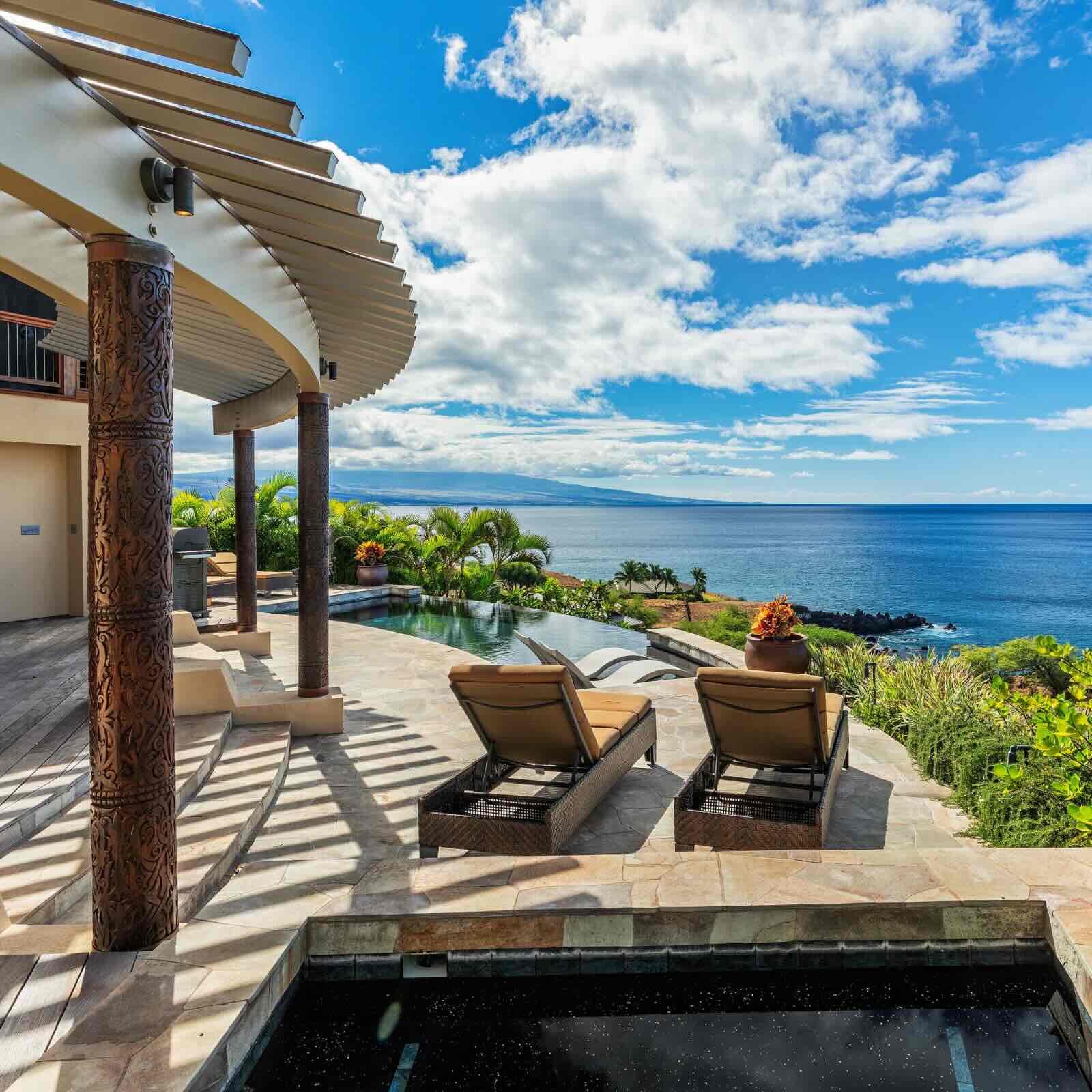 The view across the Pacific Ocean from a luxury oceanfront villa in Hawaii. The curved wooden pergola of the property is supported by decorative carved pillars, with a plunge pool and two lounge chairs visible in the foreground. An infinity pool looks out over the tranquil waters, while lush green palm trees and tropical foliage line the property edge. 