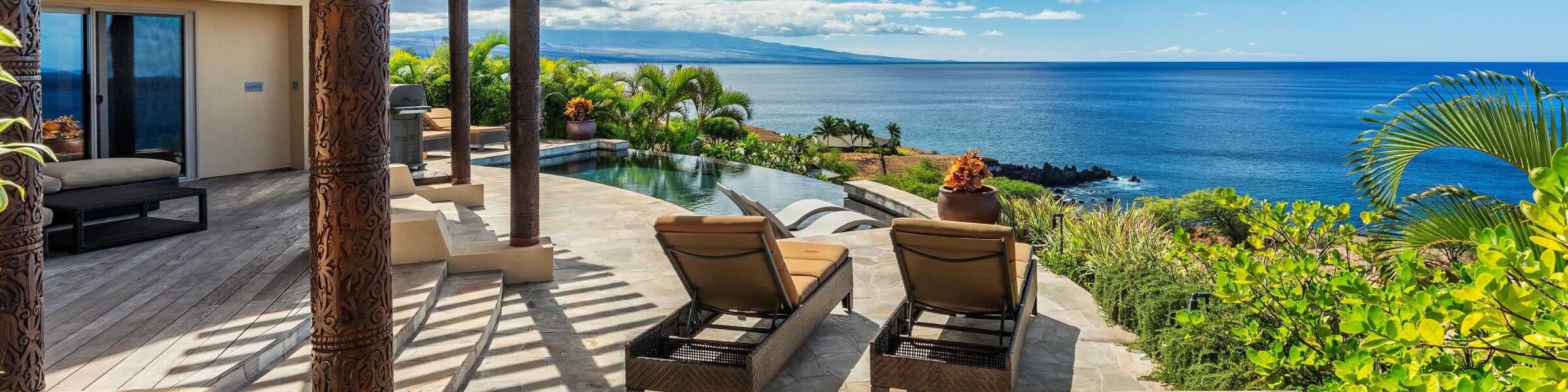 The view across the Pacific Ocean from a luxury oceanfront villa in Hawaii. The curved wooden pergola of the property is supported by decorative carved pillars, with a plunge pool and two lounge chairs visible in the foreground. An infinity pool looks out over the tranquil waters, while lush green palm trees and tropical foliage line the property edge. 