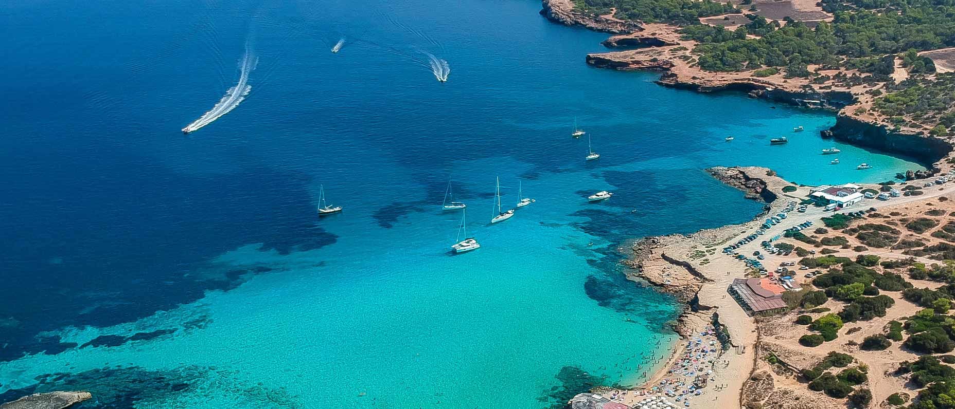 Aerial view of a turquoise beach cove with anchored boats, sunbathers and rocky, tree-lined coastline.