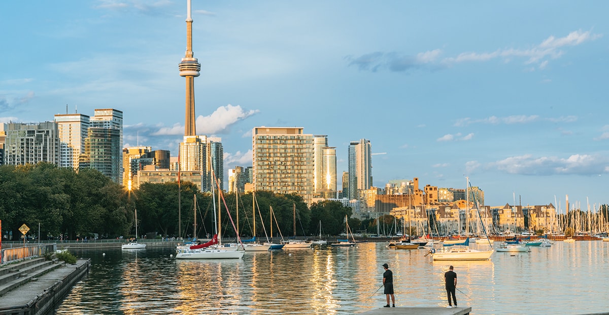 Toronto waterfront at sunset, with the CN Tower above downtown buildings and sailboats moored in the harbor as two people stand by the water’s edge.
