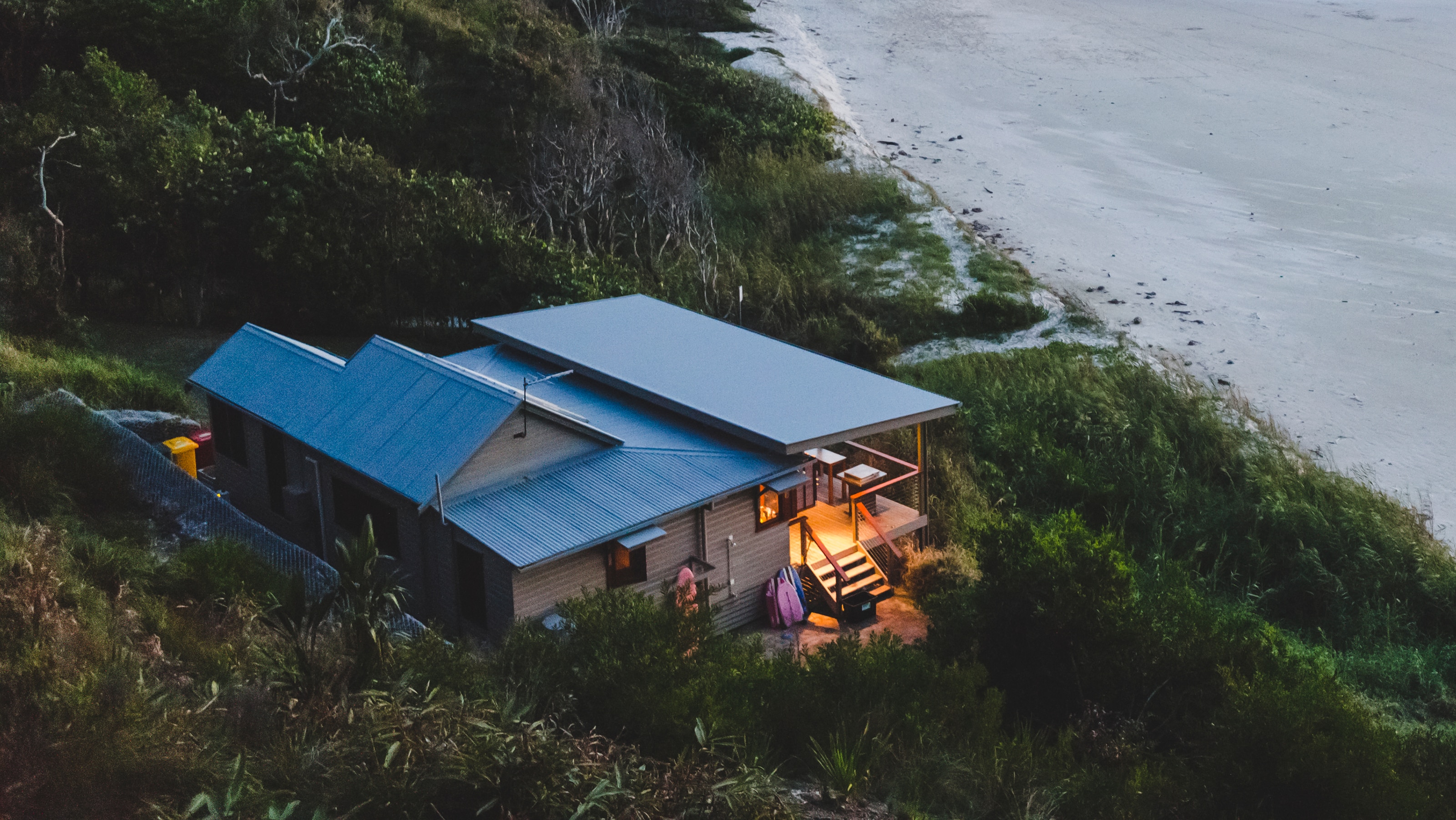 View from above of a property at dusk, with a light on over the porch and balcony, with people walking along the Australian beach and shoreline in Byron Bay. NSW.