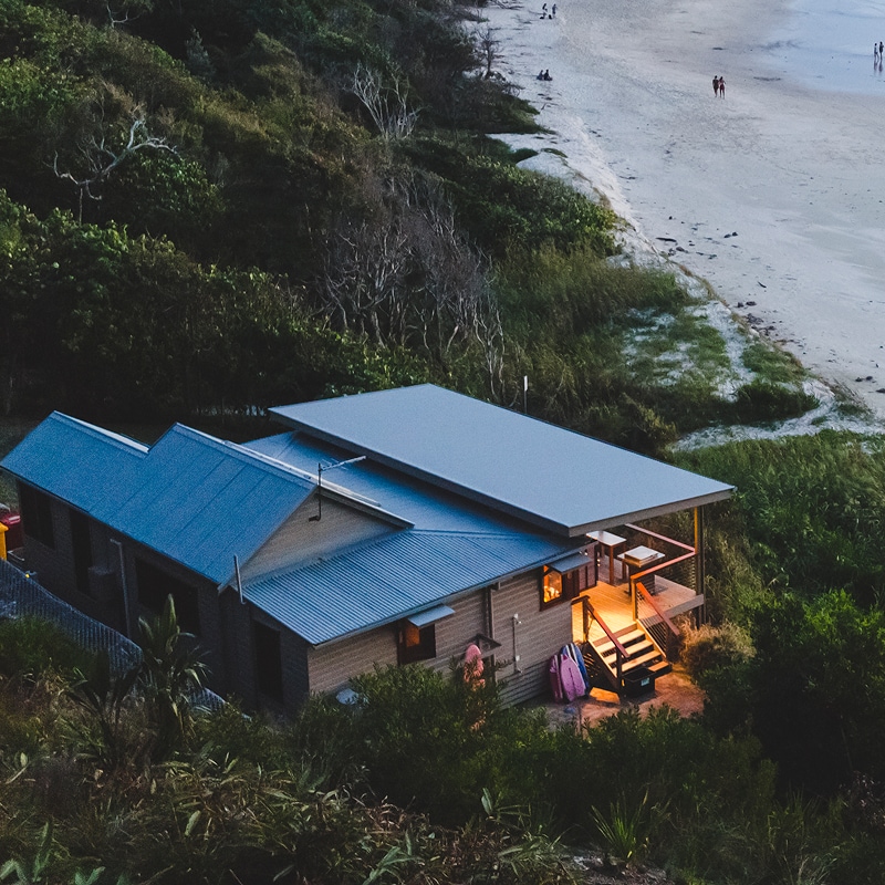 View from above of a property at dusk, with a light on over the porch and balcony, with people walking along the Australian beach and shoreline in Byron Bay. NSW.