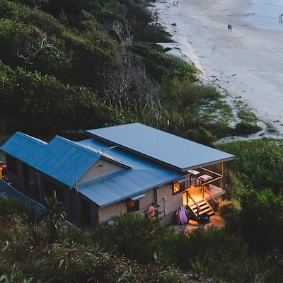 View from above of a property at dusk, with a light on over the porch and balcony, with people walking along the Australian beach and shoreline in Byron Bay. NSW.