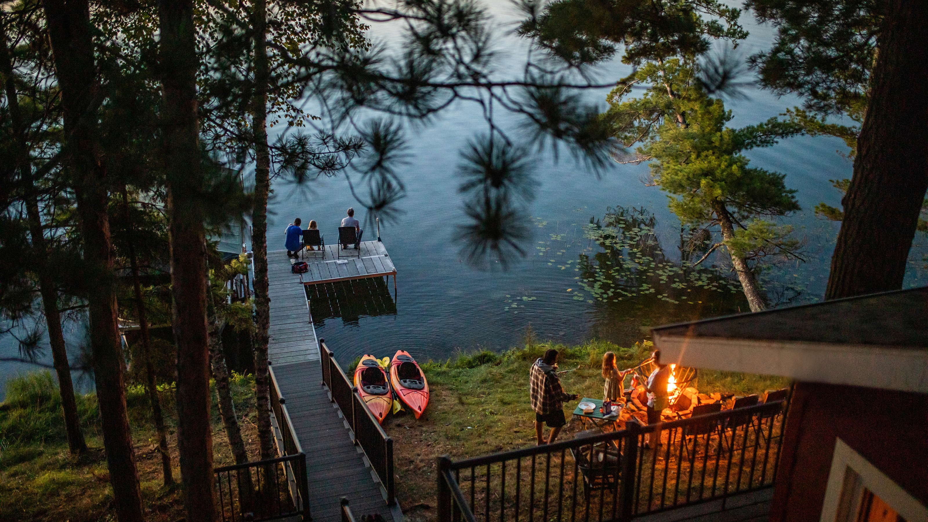 Lakeside vacation rental at dusk with a dock, kayaks, and guests relaxing by the water and around a fire pit