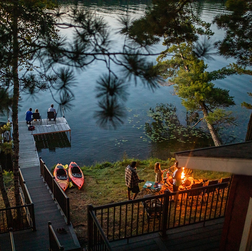 Location de vacances au bord d’un lac au crépuscule, avec un quai, des kayaks et des voyageurs qui se détendent près de l’eau et autour d’un foyer extérieur.