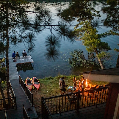 Location de vacances au bord d’un lac au crépuscule, avec un quai, des kayaks et des voyageurs qui se détendent près de l’eau et autour d’un foyer extérieur.