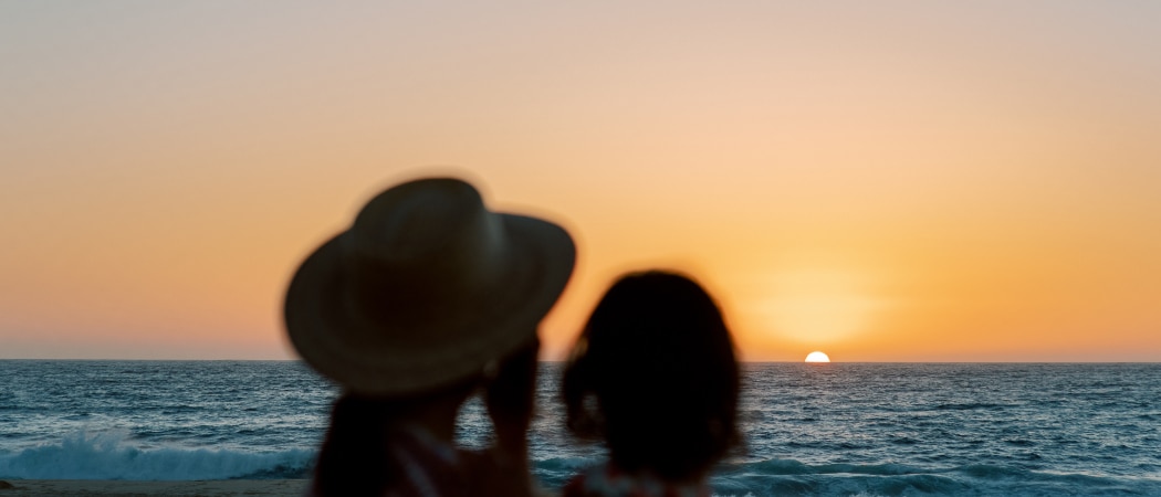 An adult wearing a sunhat holds a child as they look out at the sunset from a beach.