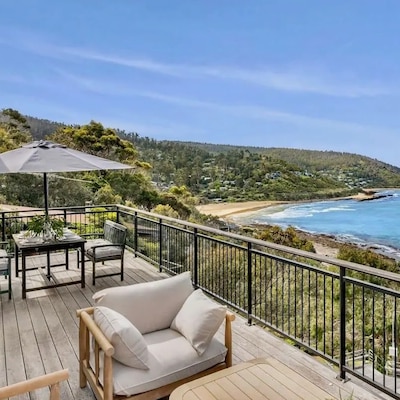 A wooden deck with chairs and a table with a grey umbrella faces a tree-covered beach and an aqua ocean.