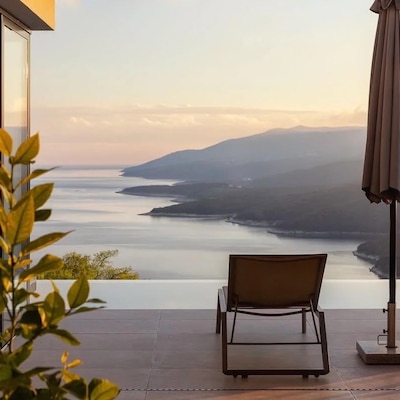 A patio with chairs and an umbrella overlooks an infinity pool facing an ocean and green hills. The sun peeks over the hills.