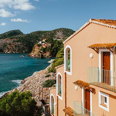 Eine sonnenbeschienene Villa am Meer blickt auf klares blaues Wasser und raue Küstenhügel.