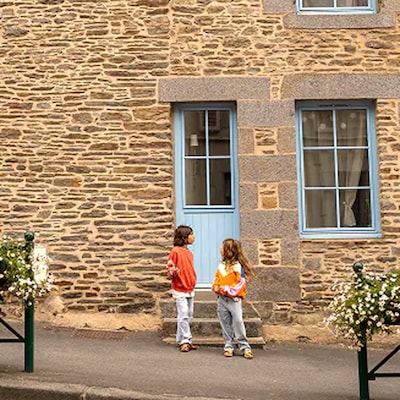 Deux enfants discutent dans une rue calme devant une maison en pierre rustique aux portes et fenêtres bleu vif.