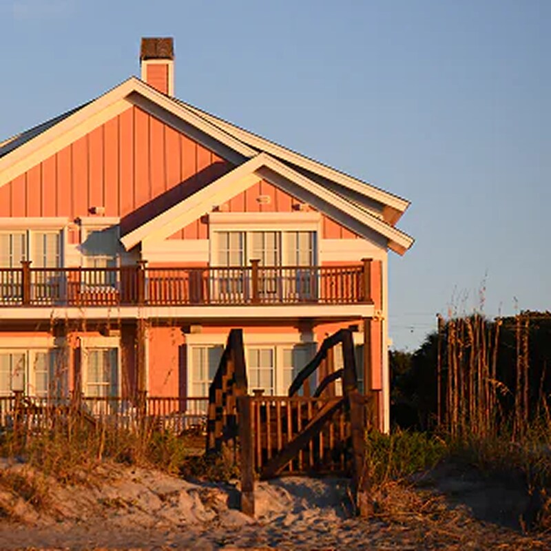 An inviting beach house glows in warm sunlight behind sandy dunes and sea grass.