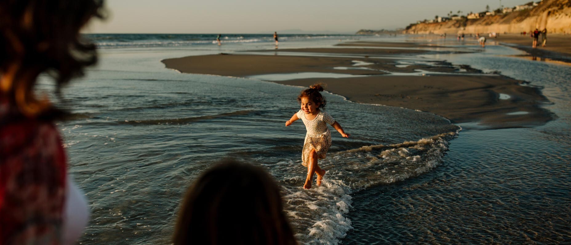 A view of an ocean coast. A child runs through the surf toward two people. Behind the child are sandbars and people walking.