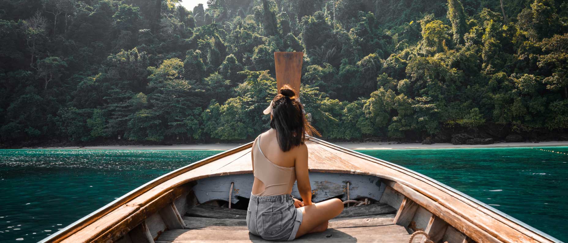 An adult sits at the front of a wooden boat while looking at dark aqua water, a sandy shore, and a sunlit forest.