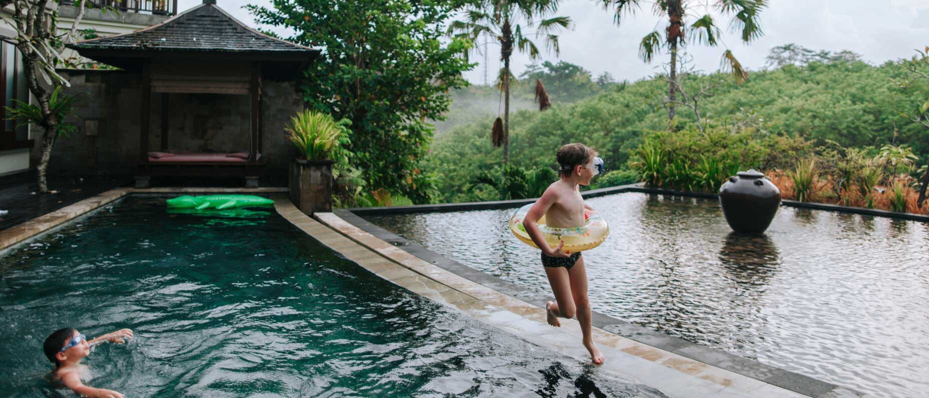 A child wearing an inflatable ring plays around a large pool at a tropical villa.