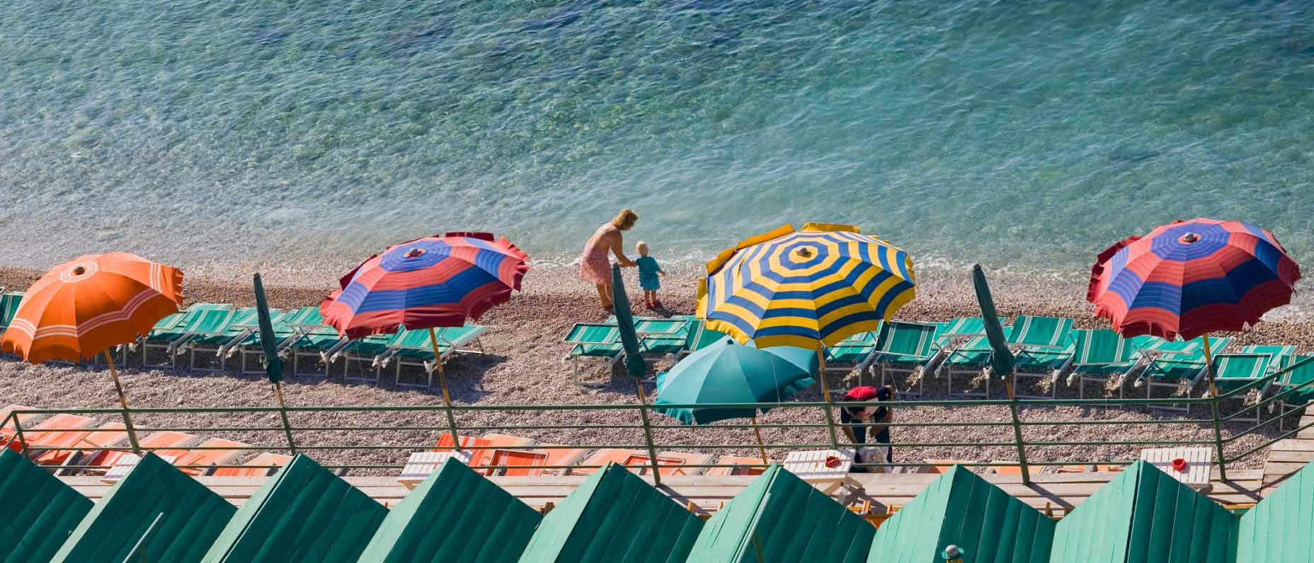 Eine Person steht am Strand am Wasserrand und hält die Hand eines Kindes, um sie herum stehen bunte Sonnenschirme. 