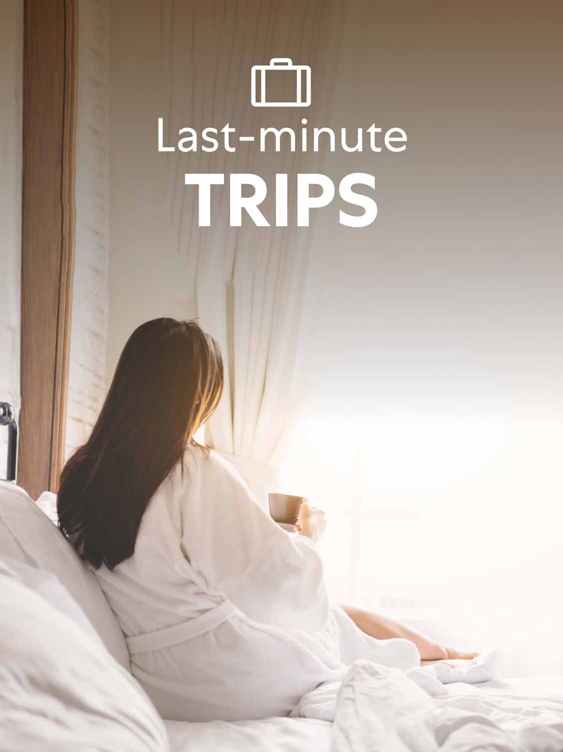 A woman in a bathrobe sits on the side of her hotel bed - drinking coffee and looking out the window. A superimposed title reads: Last-minute trips.