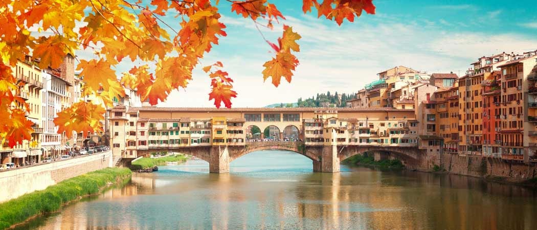 A covered foot bridge crossing a canal in Florence, Italy. A tree with Fall colored leaves hangs in the foreground.