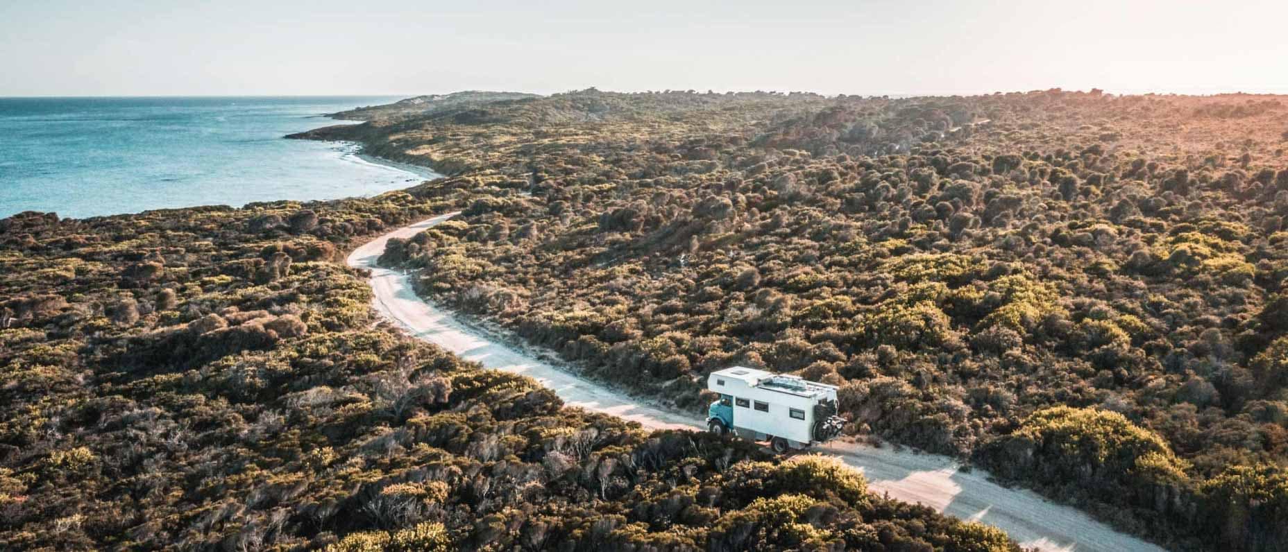 Aerial view of a single road winding through a rocky bush landscape. A vintage campervan drives towards a beach in the distance.