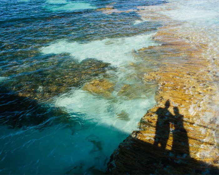 The sun is shining across shallow ocean pools and casts the shadows of two people looking at the water. 