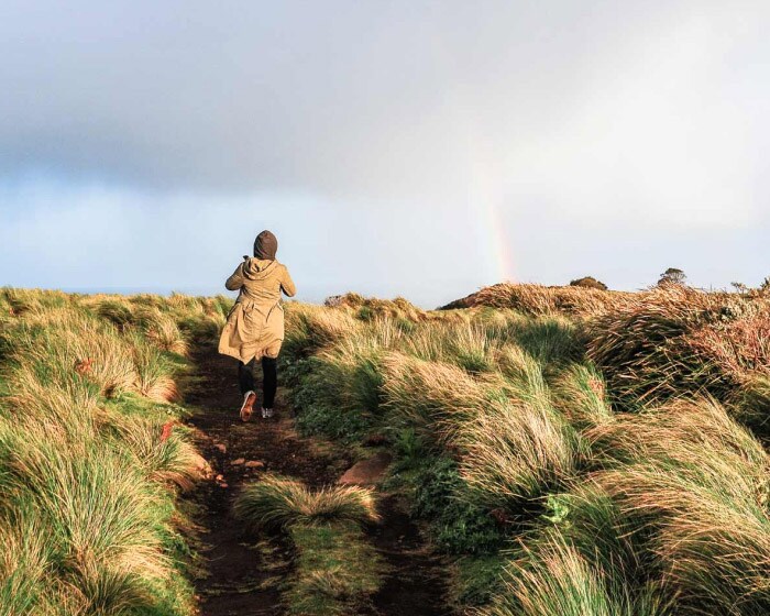 A person wearing a long coat hikes across a shrubby field walking towards a rainbow.