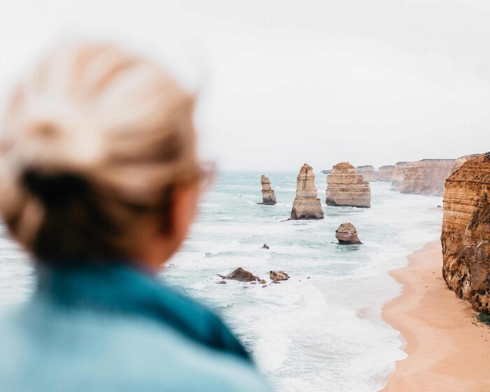 A person in a blue coat looks out at the 12 Apostles rock formations and cliffs on the Great Ocean Road. 