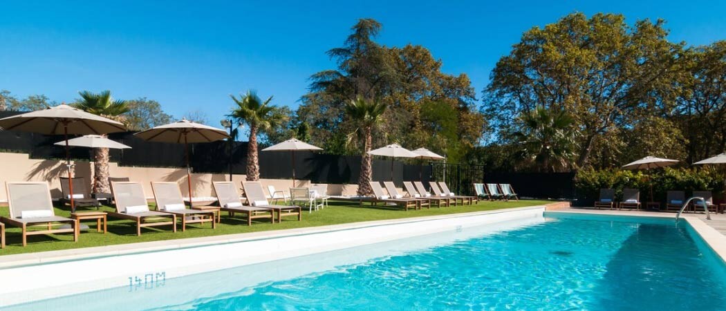 Exterior view of white umbrellas and timber lounge chairs beside a swimming pool at the Hôtel SPA de Fontcaude.