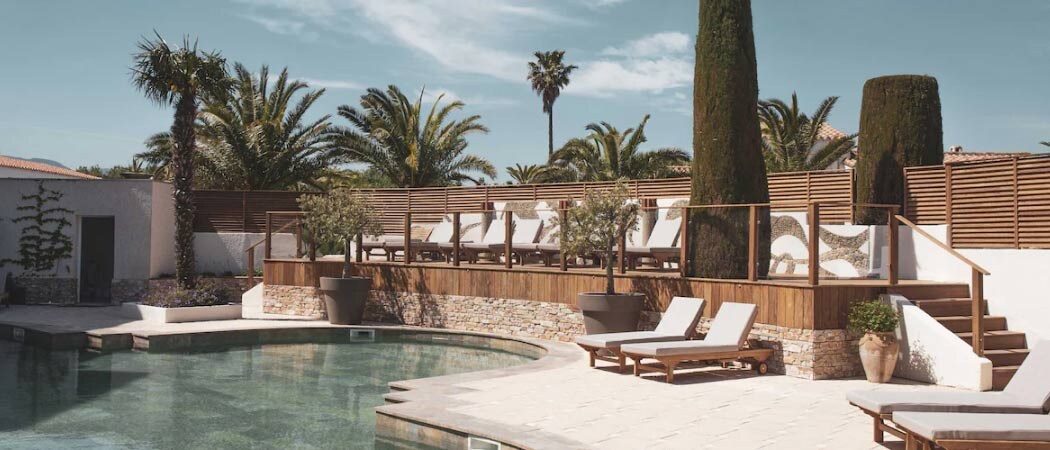 Exterior view of white lounge chairs underneath palm trees beside a swimming pool at the L’ Île de la Lagune Thalasso & Spa