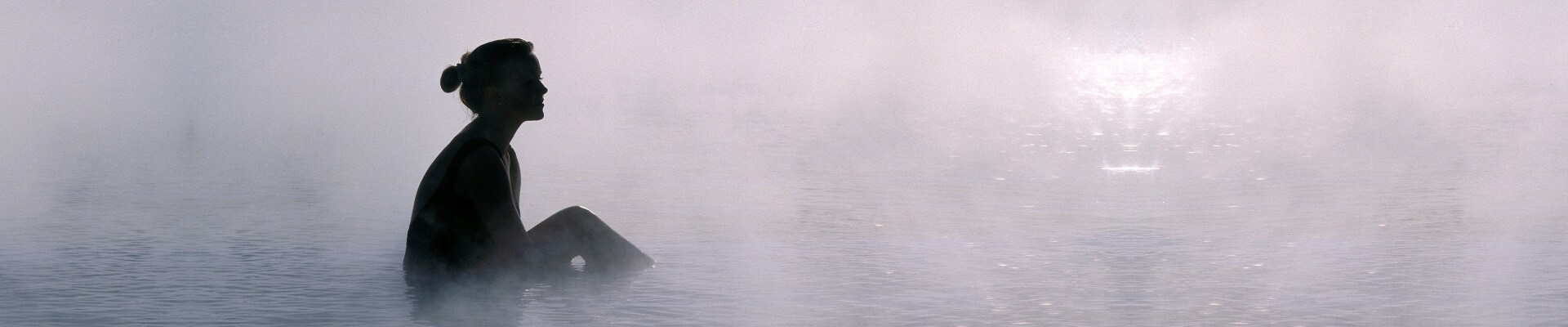 Steam rises from a pool of water and a person sits in the middle looking off into the distance.