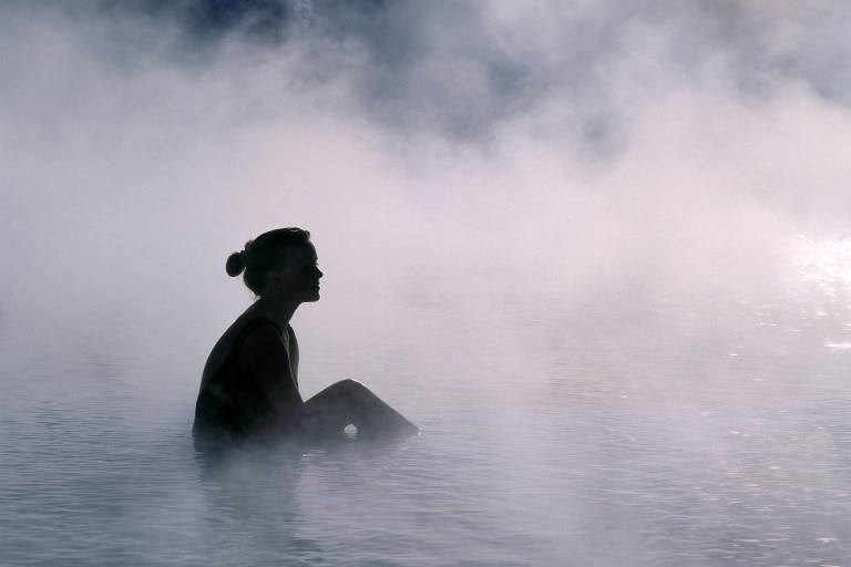 Steam rises from a pool of water and a person sits in the middle looking off into the distance.