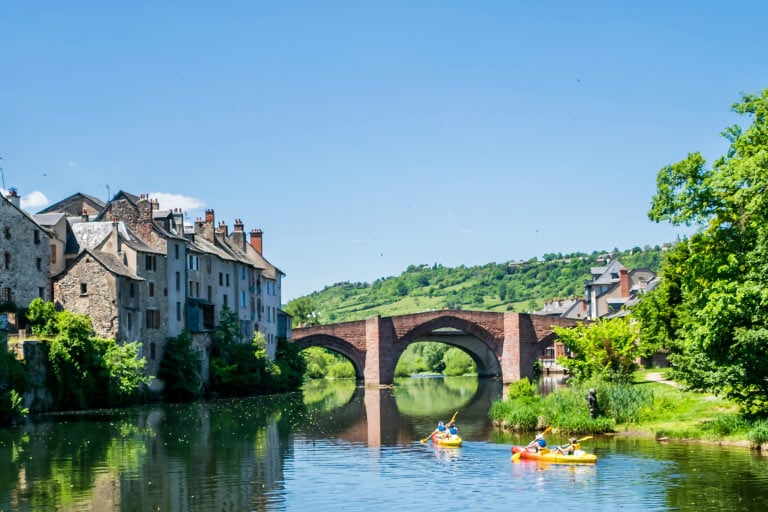 Two yellow kayaks float on a river in Espalion, France. A bridge is in the background, buildings and trees line the banks.