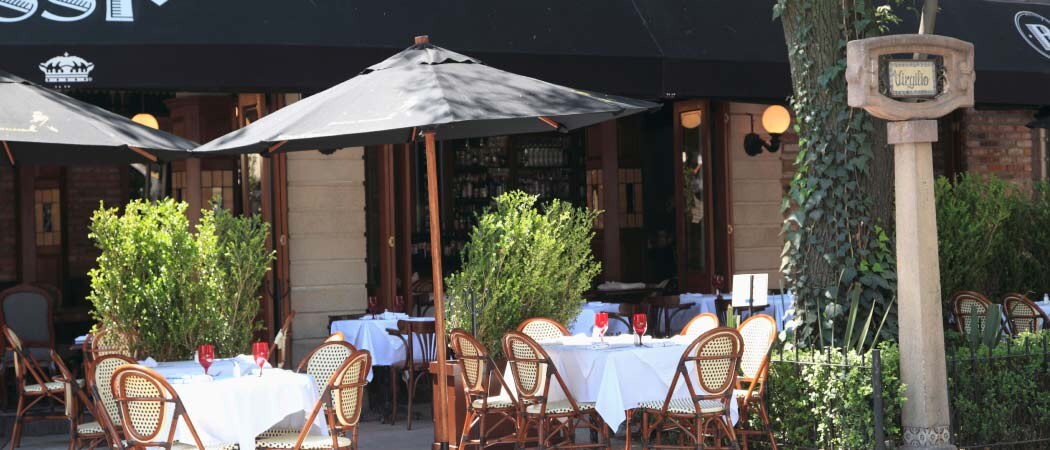 An exterior view of a cafe. A group of tables and chairs are arranged in the sun underneath large, black umbrellas.