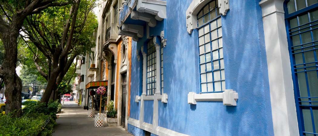 Exterior view of a tree-lined street with a row of colorful two-story houses with small balconies.