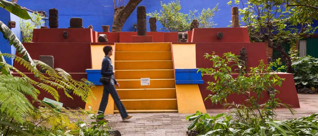 A person walks in front of a red, blue, and yellow structure covered in sculptures at the Frida Kahlo museum garden.